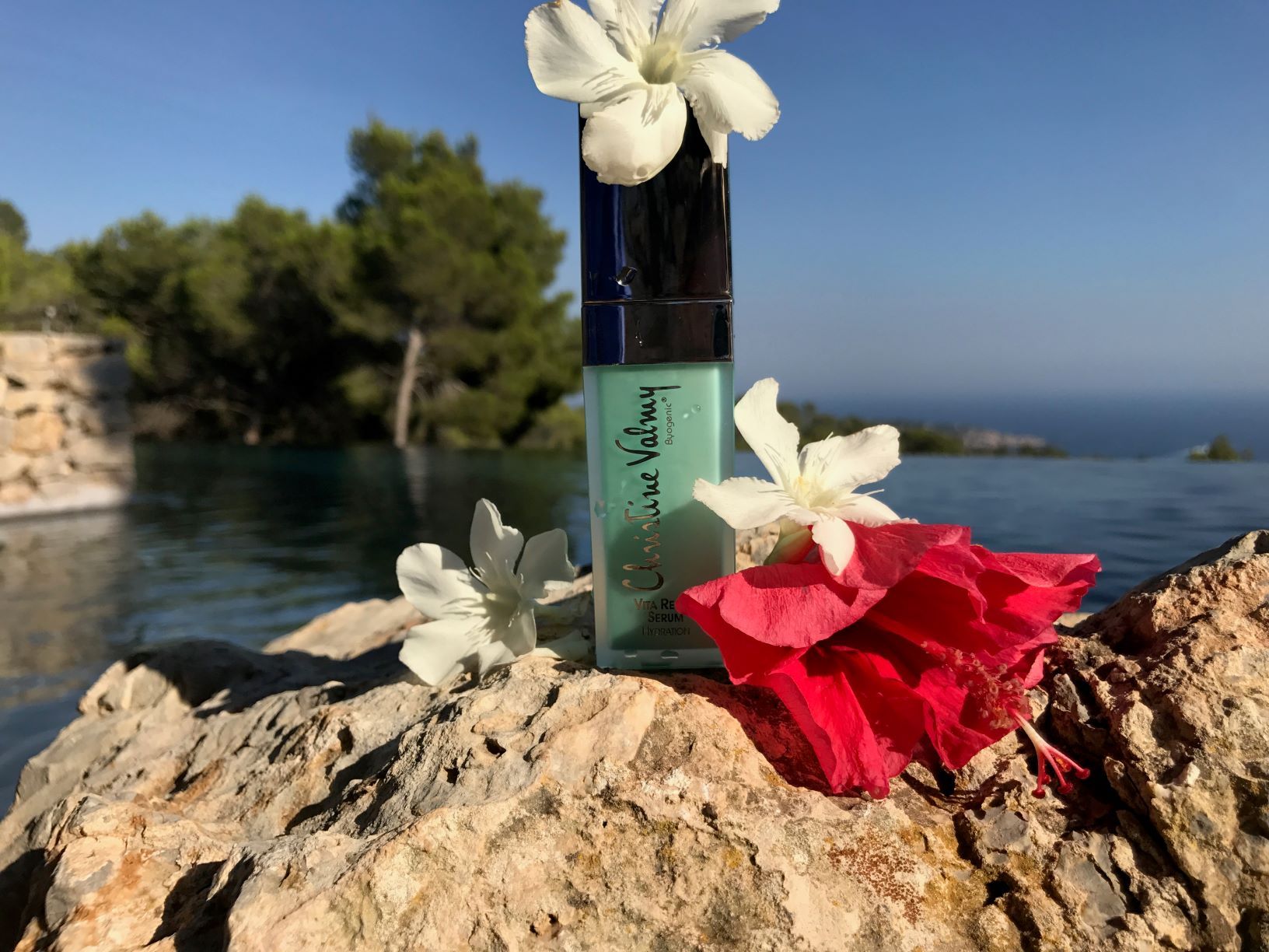 Christine Valmy Skin Care Product on rocks, surrounded by flowers, in front of a body of water.