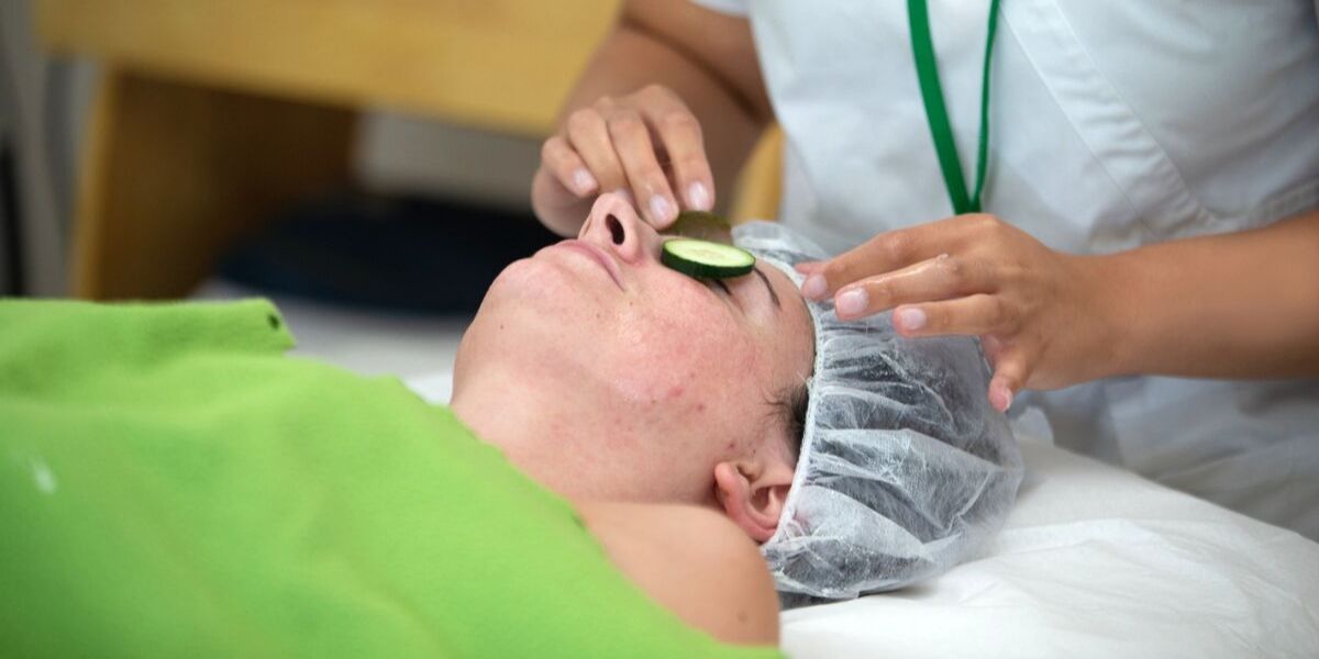 Skin care professional performing a facial on a client with cucumbers on their eyes.